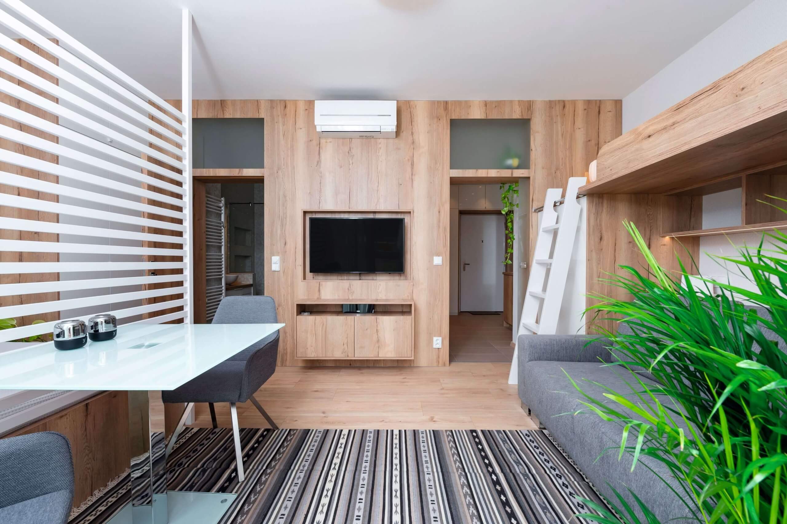 Heller Wohnraum mit integrierter TV-Wand in Holzoptik. Raumteiler mit weißen Lamellen sorgt für Struktur. Graue Sitzmöbel, Leiter zum Hochbett und viele Pflanzen schaffen ein modernes, gemütliches Ambiente.
