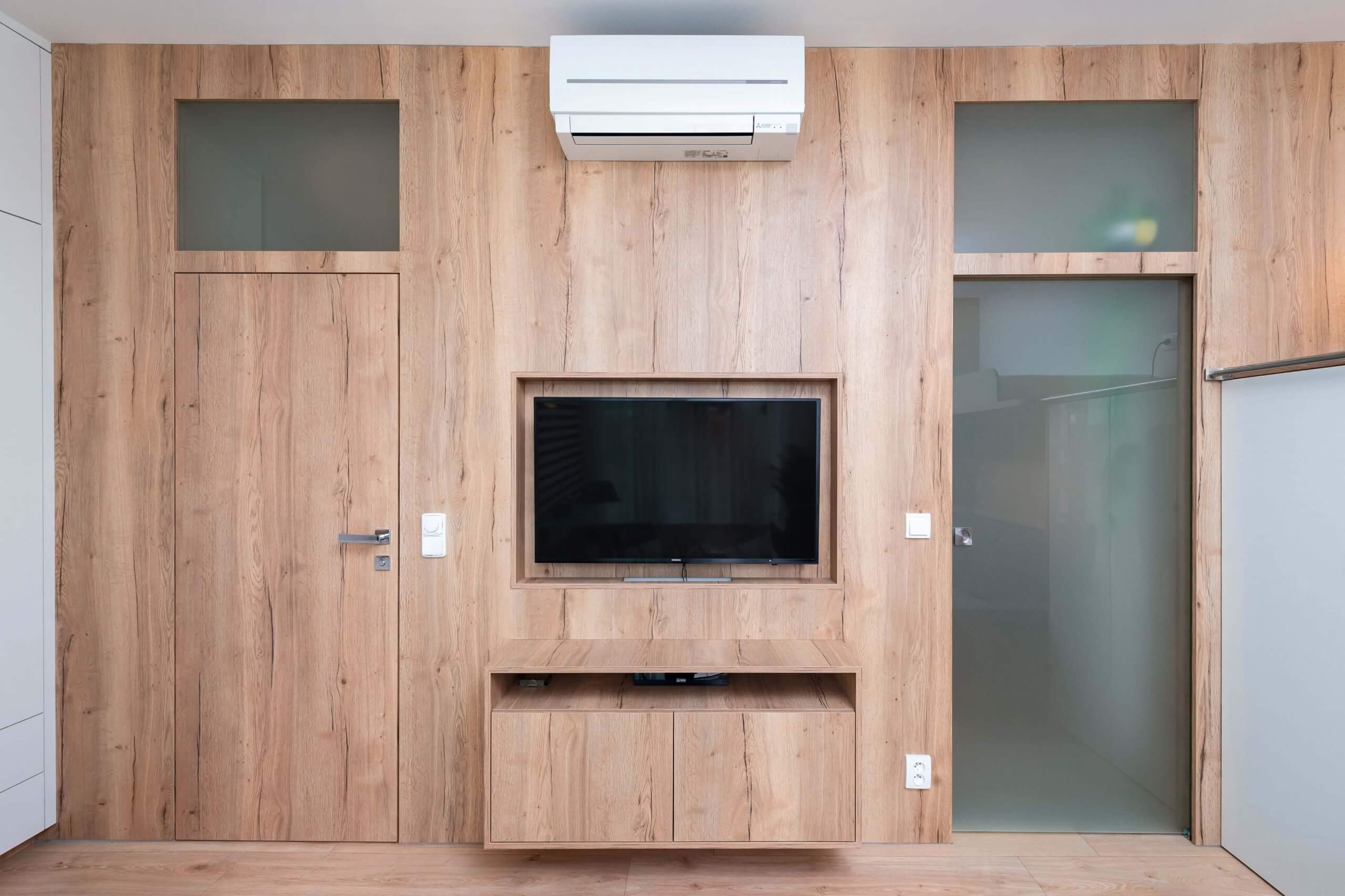 Moderne Medienwand mit durchgängiger Holzverkleidung in natürlicher Optik. Integrierter Fernseher und Unterschrank sorgen für Ordnung. Zwei Türen mit satiniertem Glasaufsatz und eine Klimaanlage runden das funktionale Design ab.