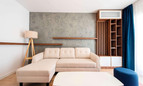 Helles, modernes Wohnzimmer mit beigem Ecksofa, rustikaler Betonwand, Holzregalen und blauen Akzenten – stilvolle und gemütliche Einrichtung.