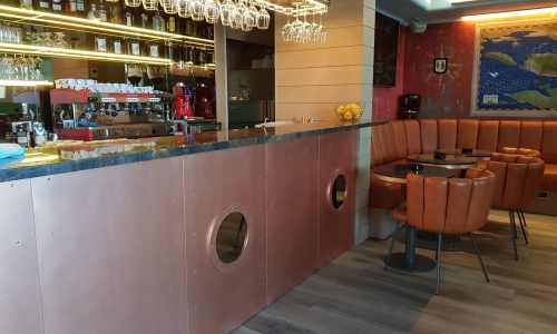Stilvolle Bar mit messingfarbenem Tresen, integrierten Bullaugen und gemütlicher Leder-Sitzbank – einladendes Ambiente mit maritimer Note.