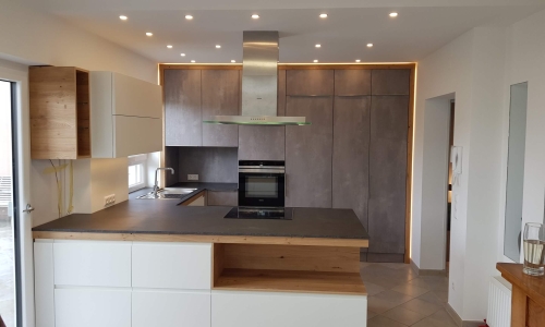 Moderne Küche mit zentraler Kochinsel, grauen Hochschränken, weißen Fronten und stilvollen Holzelementen – elegante LED-Beleuchtung rundet das Design ab.