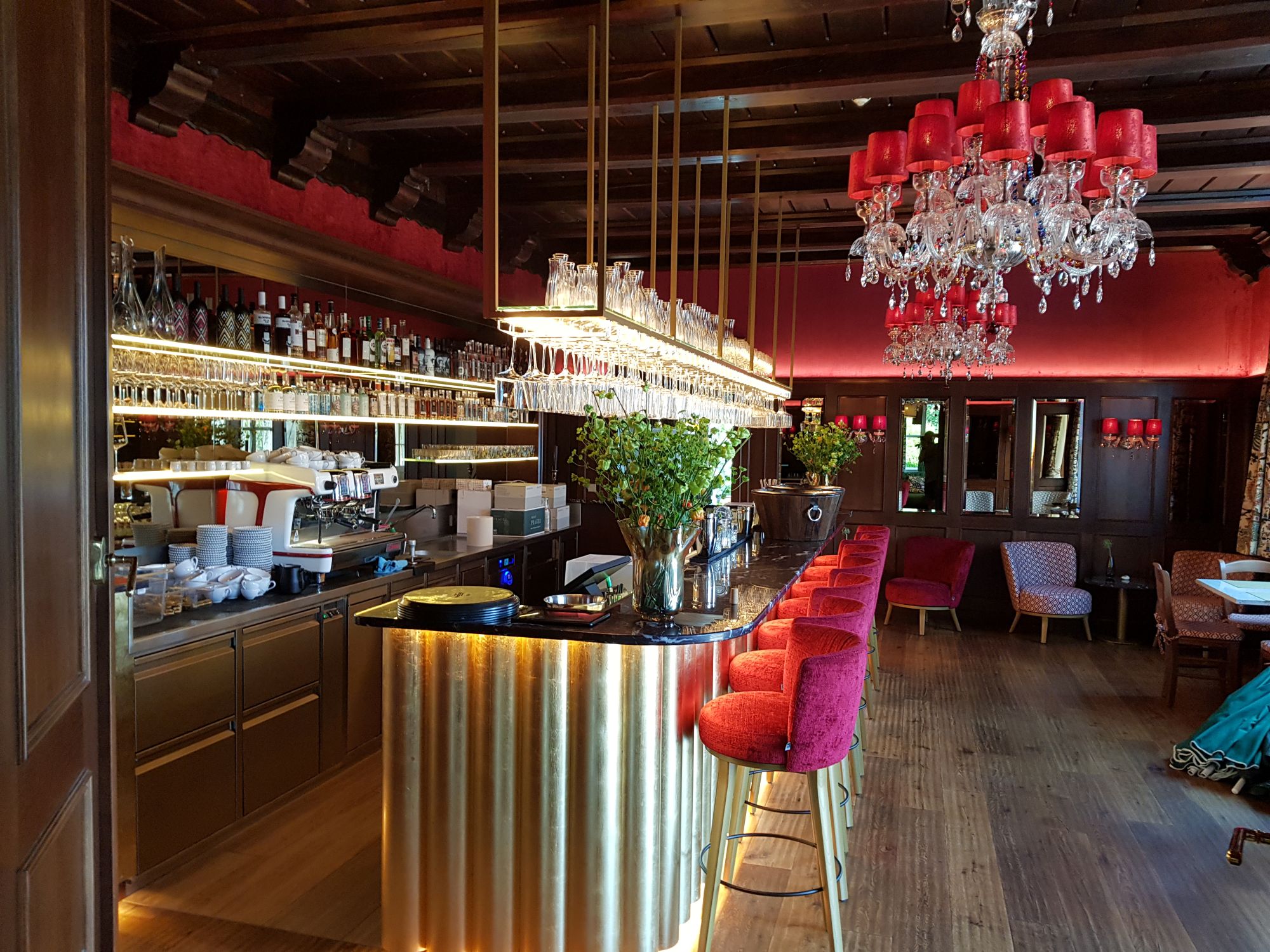 Edle Bar mit goldverkleideter Theke, roten Samthockern, opulentem Kronleuchter und stilvoller Beleuchtung – ideal für gehobene Abende.